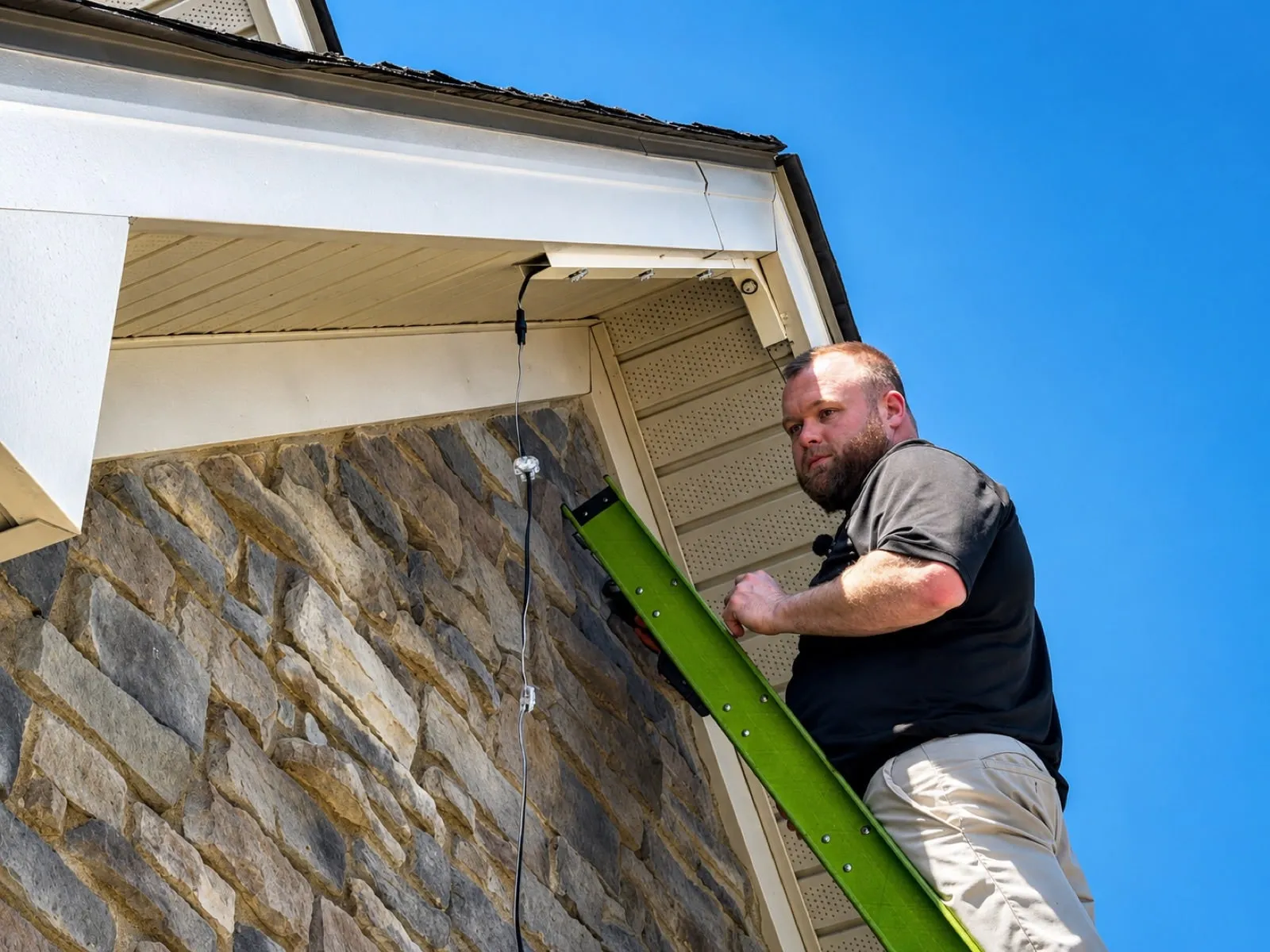 Installer on a ladder fastening permanent light track under the soffit of a stone-front home